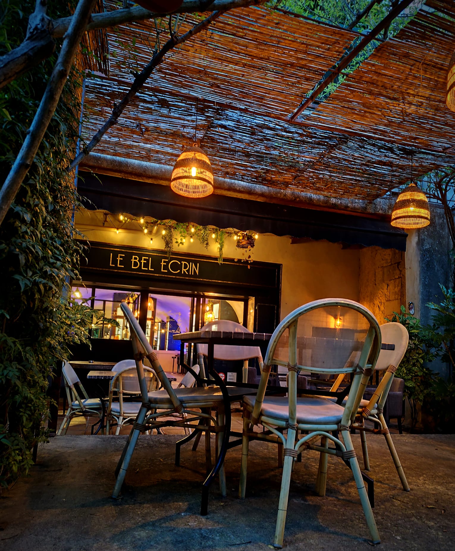 Le patio du Bel Écrin à la tombée de la nuit : tables en terrasse, lanternes en osier et enseigne éclairée